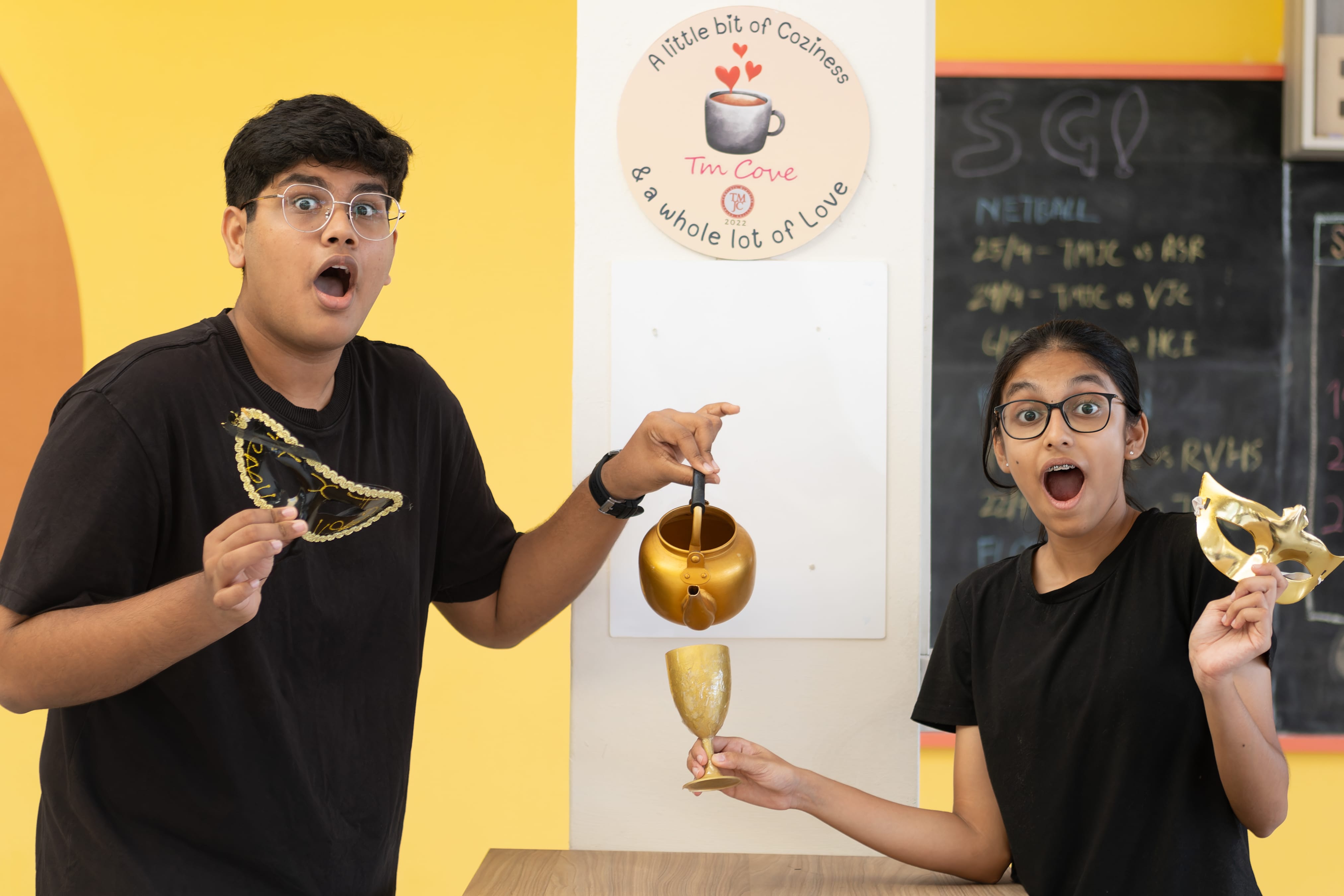 Two students in black T-shirts stand on either side of a white pillar with exaggerated expressions of surprise, each holding theatrical gold props including masks, a teapot, and a goblet.